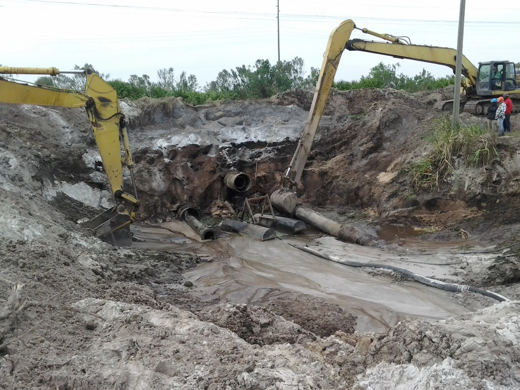 Two excavators working in a deep, muddy excavation with exposed pipe