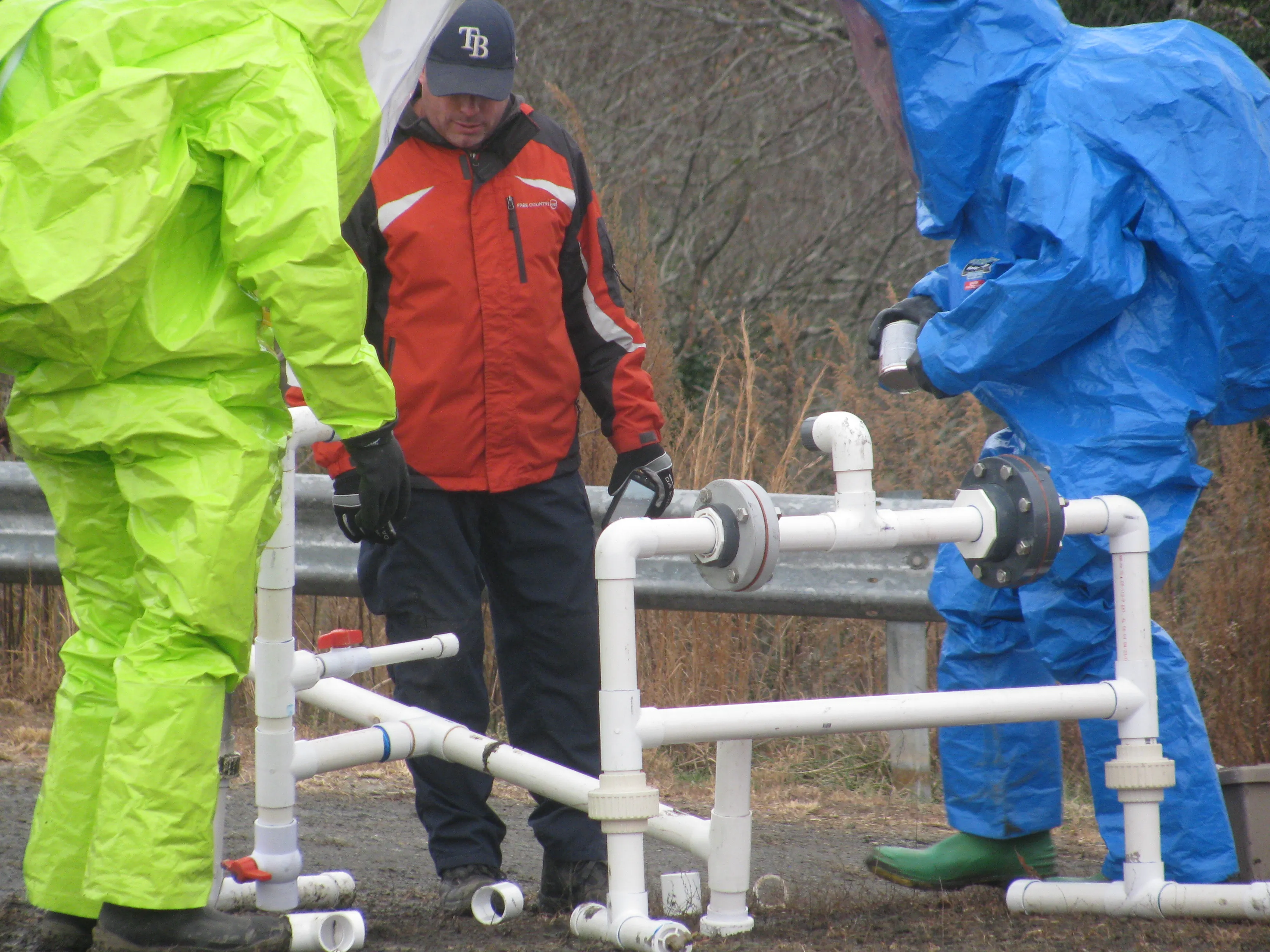 Trainees in chemical protective suits working a leaking valve prop while an instructor watches